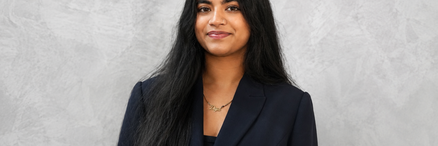 Diya Kulkarni, a young woman with long dark hair, wearing a black blazer and a gold necklace, smiles in front of a gray background.