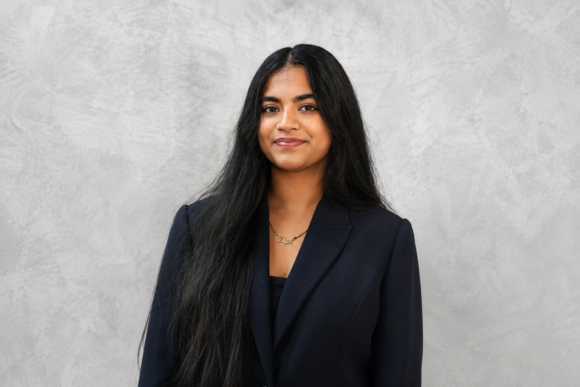 Diya Kulkarni, a young woman with long dark hair, wearing a black blazer and a gold necklace, smiles in front of a gray background.