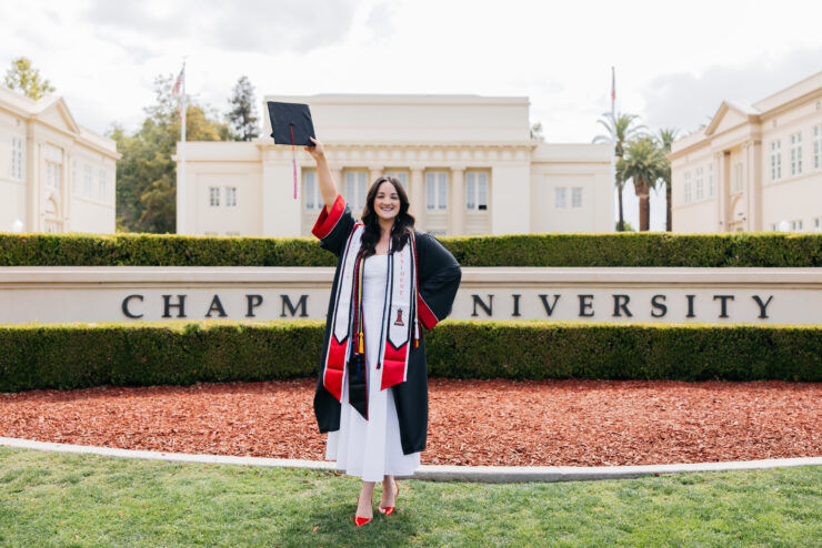 Olivia Marr wears a white dress and graduation regalia as she holds her mortarboard cap in the air while standing in front of the Chapman University sign.