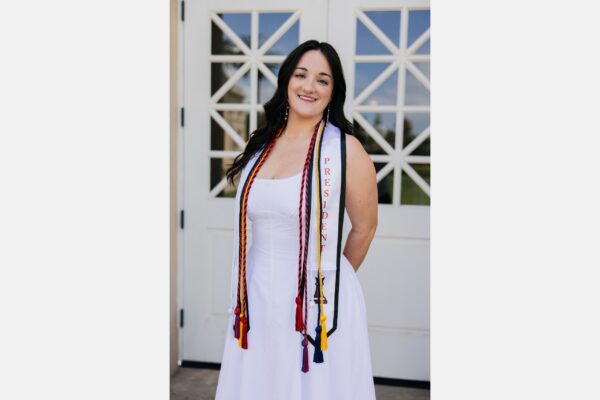 Olivia Marr, a young woman in a white dress, stands in front of the fenestra windows of Memorial Hall wearing multiple graduation stoles.