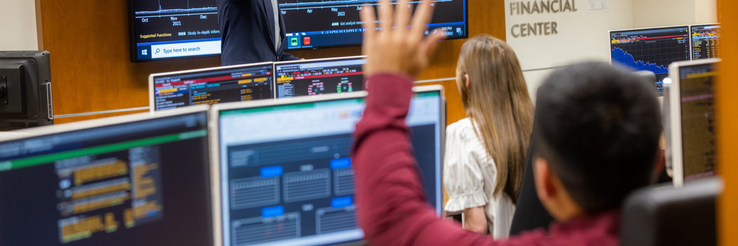 man standing in front of a screen displaying stock market information pointing towards a seated male student with his hand raised.