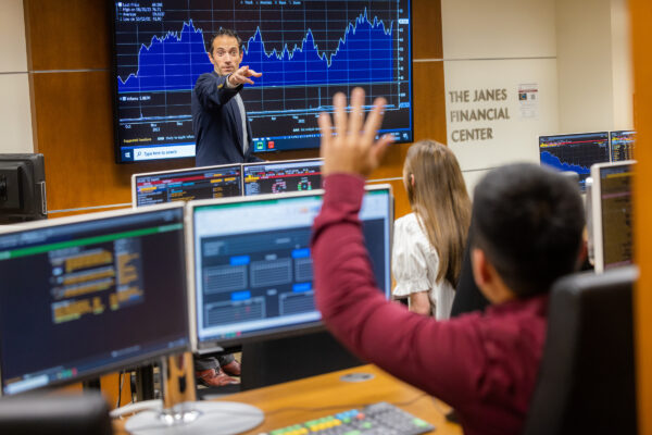 man standing in front of a screen displaying stock market information pointing towards a seated male student with his hand raised.