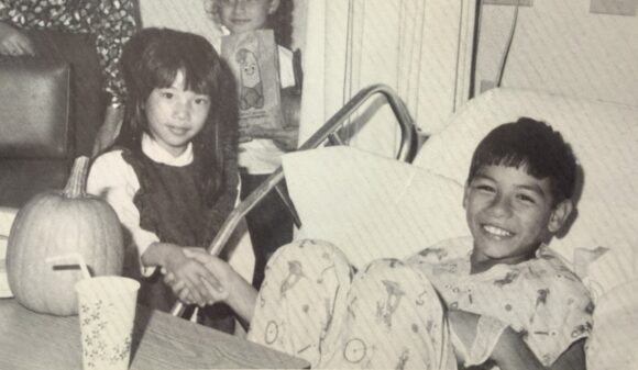 Black and white photo of a young Grace Fong shaking hands with a smiling boy in a hospital bed, a pumpkin on the table beside them.
