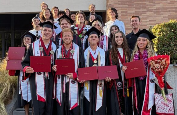 A group of Chapman University track and field seniors in graduation caps and gowns pose together on the field at commencement. Many wear "Student Athlete" stoles, flower leis, medals, and honor cords.