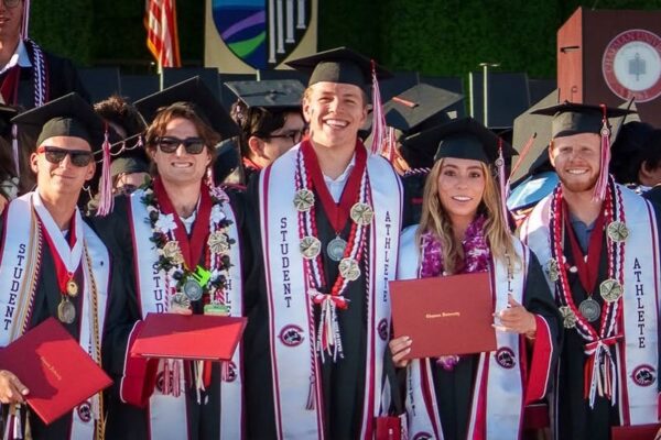 Five Chapman University graduates in caps and gowns stand together smiling on a stage at commencement. All wear "Student Athlete" stoles and medals; one holds a red Chapman University diploma cover.