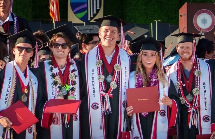 Five Chapman University graduates in caps and gowns stand together smiling on a stage at commencement. All wear "Student Athlete" stoles and medals; one holds a red Chapman University diploma cover.