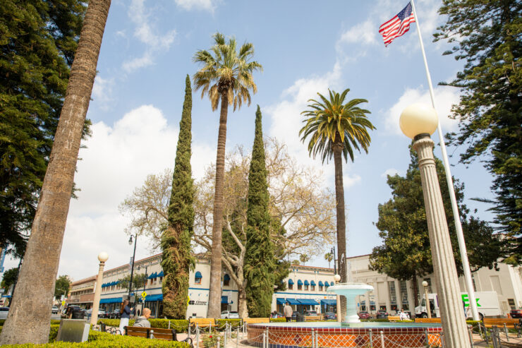 Town square scene featuring tall palm trees and other greenery surrounding a circular fountain in the middle, with people sitting on benches, historic low-rise buildings with blue awnings in the background, and an American flag on a high pole against a partly cloudy sky.