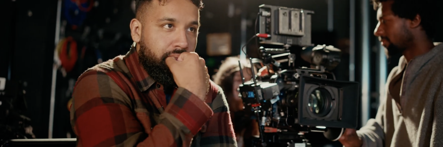 A male screenwriting student standing next to a movie camera watching a scene be filmed.