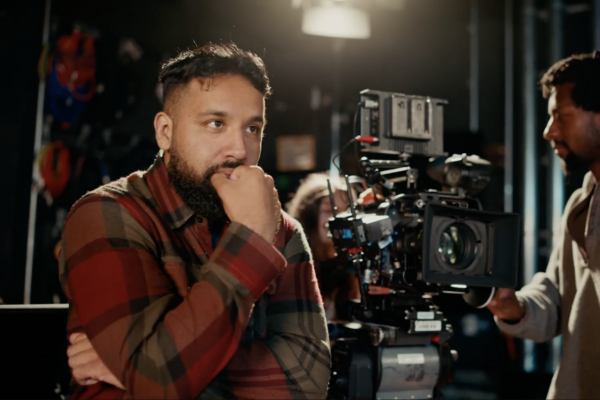 A male screenwriting student standing next to a movie camera watching a scene be filmed.