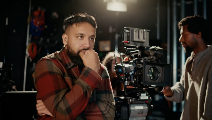 A male screenwriting student standing next to a movie camera watching a scene be filmed.
