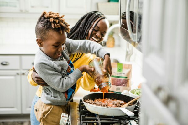An adult and young child smile as they prepare a meal together in a home kitchen, pouring tomato sauce into a skillet on the stove.