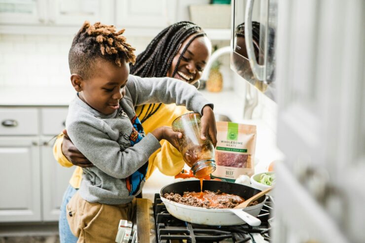 An adult and young child smile as they prepare a meal together in a home kitchen, pouring tomato sauce into a skillet on the stove.