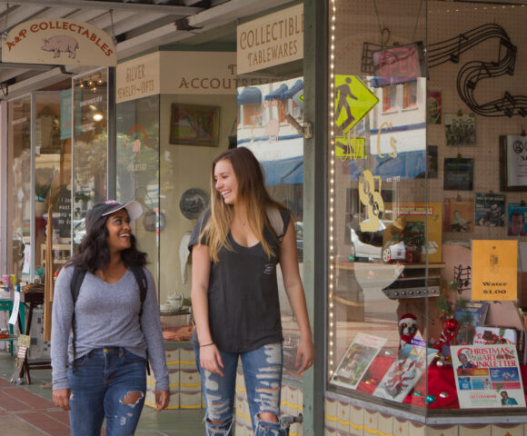 Two young women walk along a sidewalk, smiling and talking as they pass storefronts and collectible shops in a downtown shopping area.