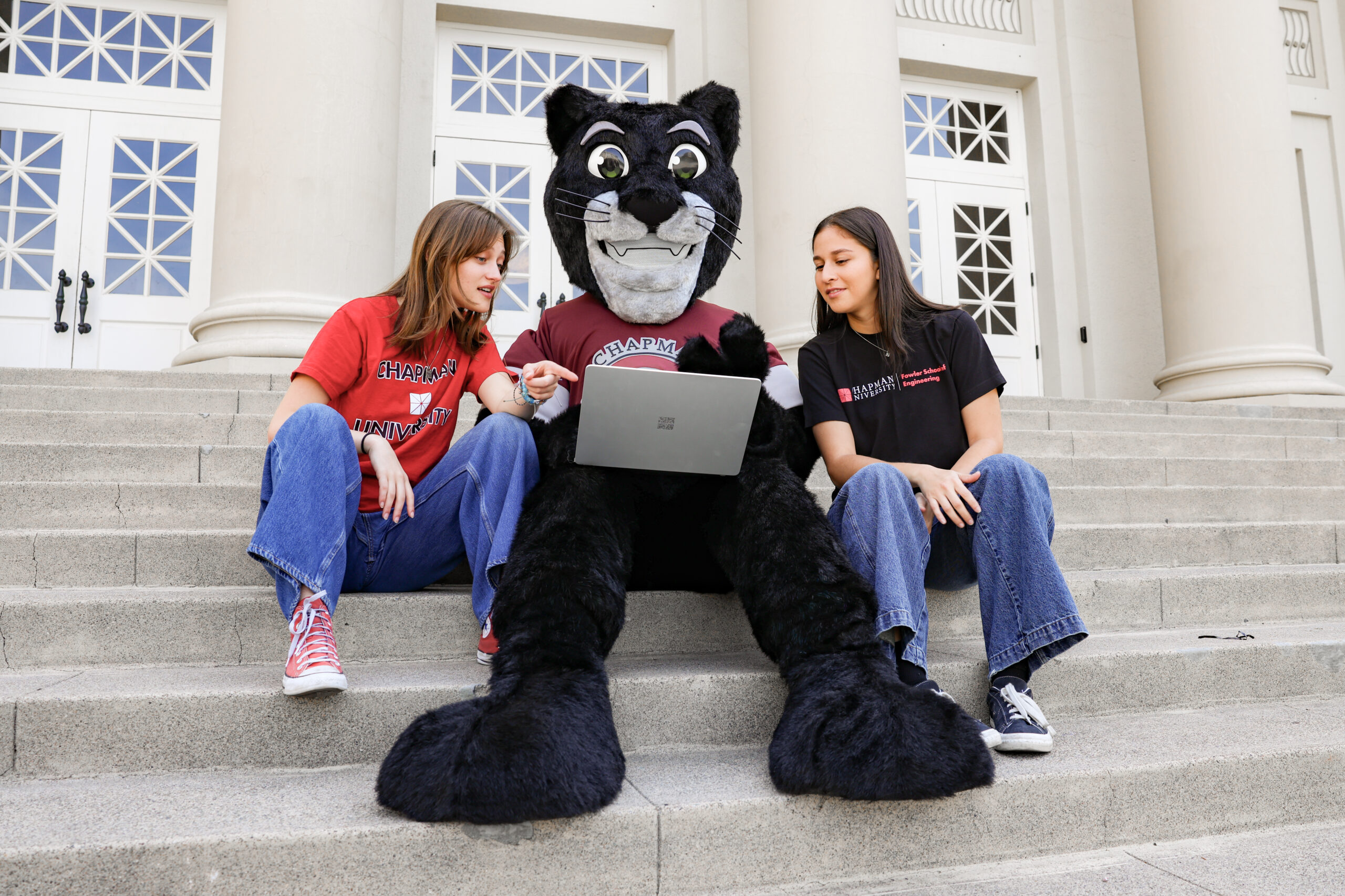 Early Action vs. Regular Decision: Two students sit with Chapman University’s Pete the Panther mascot on campus steps, looking at a laptop.