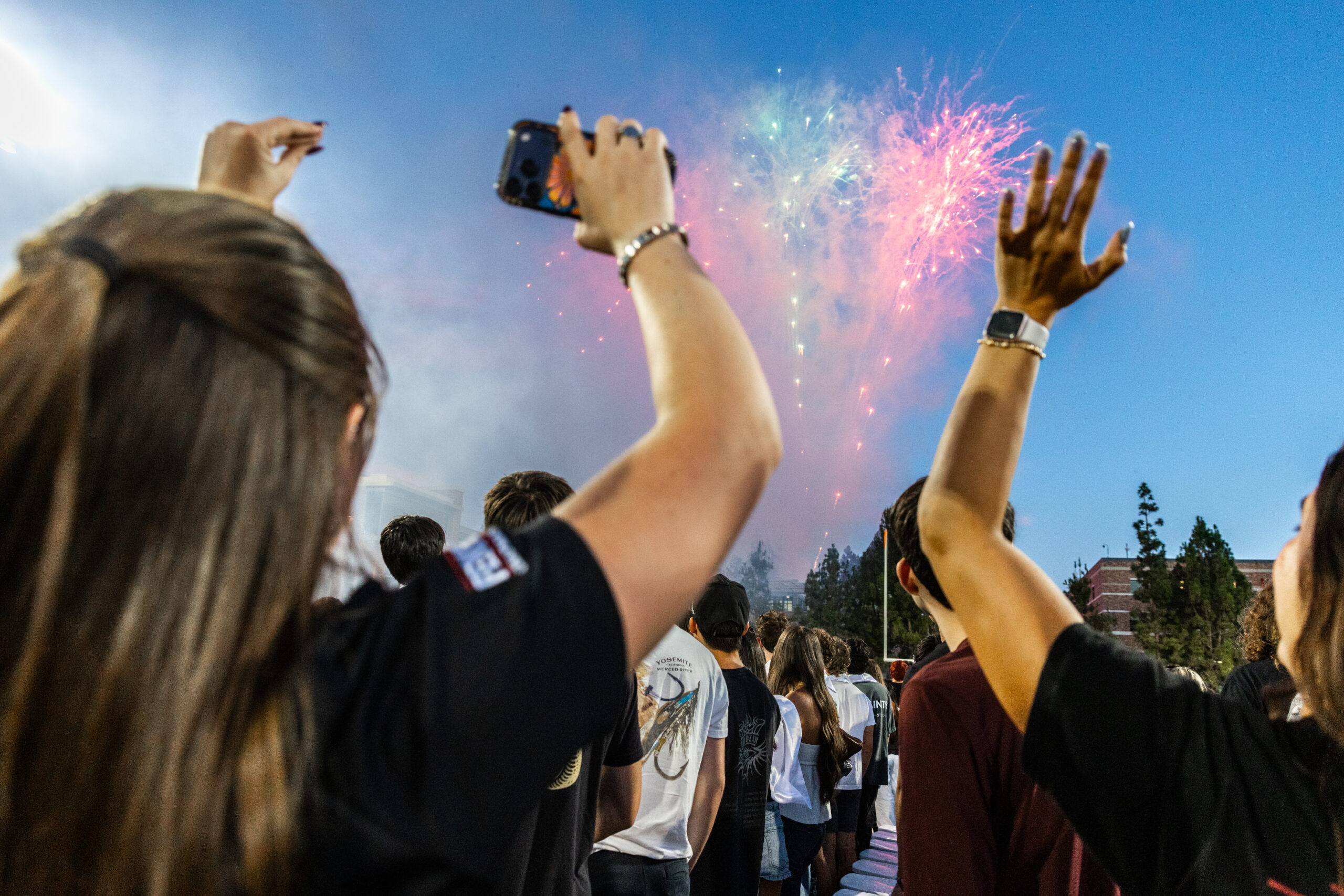 Students raise their hands and record colorful fireworks in the evening sky with a cellphone.