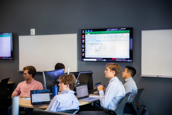 Accounting students work on laptops while a spreadsheet is displayed on a TV screen.