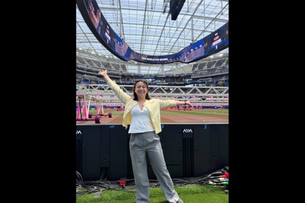 Caroline Chang stands with her arms outstretched beside the field at SoFi Stadium beneath a FIFA World Cup 2026 video board.