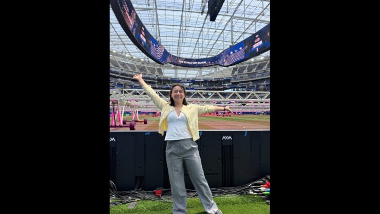 Caroline Chang stands with her arms outstretched beside the field at SoFi Stadium beneath a FIFA World Cup 2026 video board.