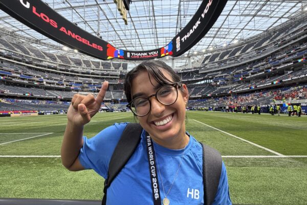 Bella Ortiz smiles and raises one hand beside the field at SoFi Stadium during the FIFA World Cup.