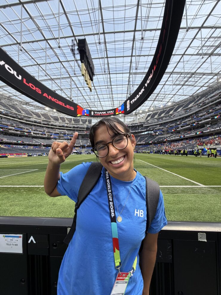 Bella Ortiz smiles and raises one hand beside the field at SoFi Stadium during the FIFA World Cup.