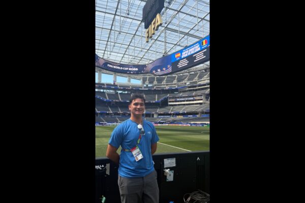 Lucas Patton smiles beside the field at SoFi Stadium beneath FIFA World Cup displays.