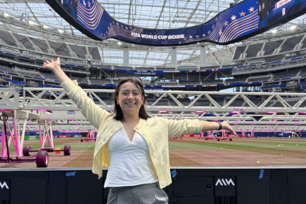 Caroline Chang stands with her arms outstretched beside the field at SoFi Stadium beneath a FIFA World Cup 2026 video board.