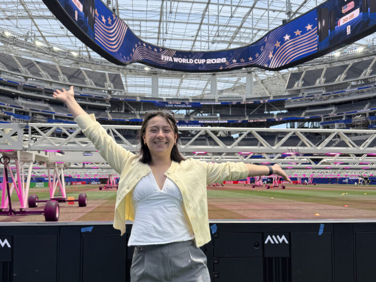 Caroline Chang stands with her arms outstretched beside the field at SoFi Stadium beneath a FIFA World Cup 2026 video board.