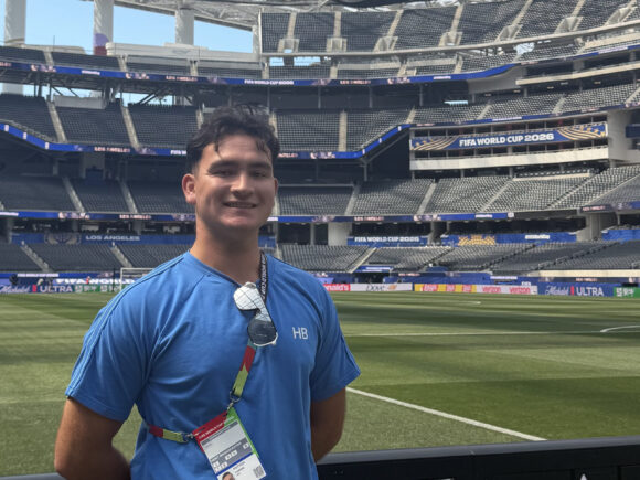 Lucas Patton smiles beside the field at SoFi Stadium beneath FIFA World Cup displays.