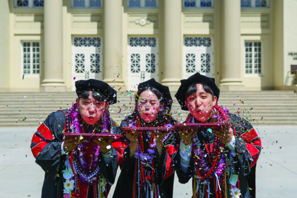 Three graduates dressed in cap and gowns blow confetti as they stand in front of Memorial Hall.