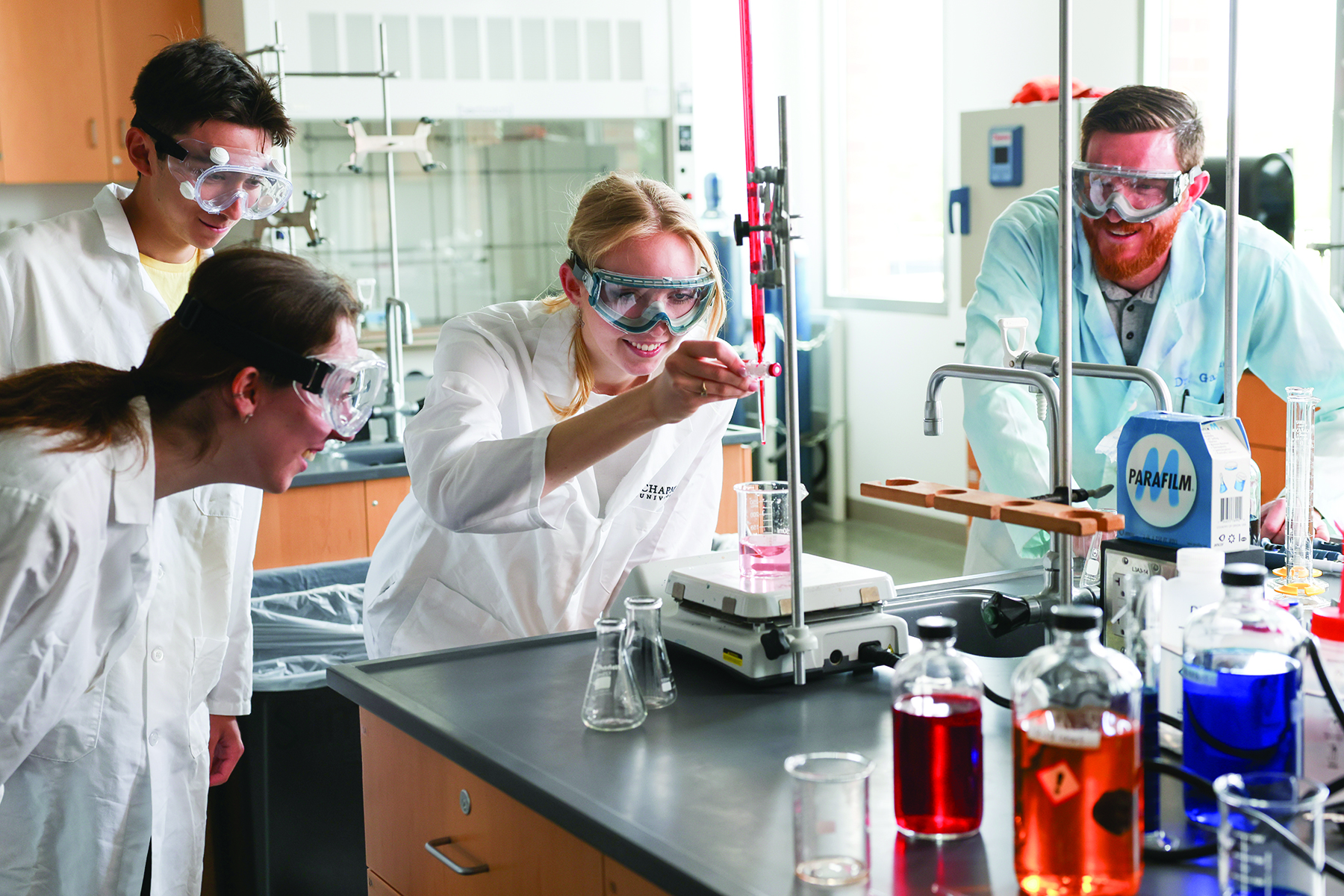Three students and a professor wearing lab coats and safety goggles conduct an experiment together in a chemistry lab.