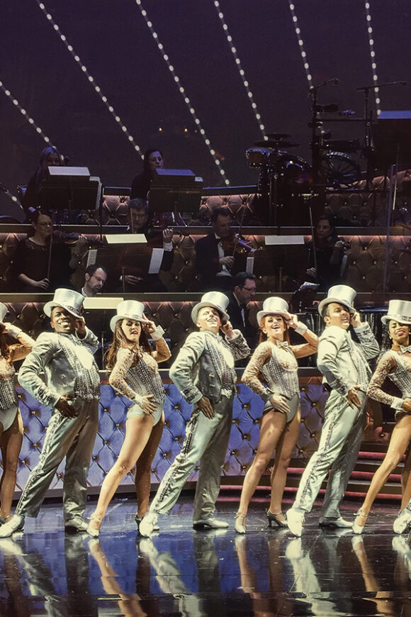 Joe Chantry performs in a silver top hat and costume with an ensemble of dancers as an orchestra plays behind them.