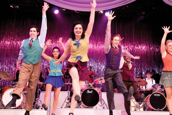 Joe Chantry and an ensemble of dancers perform a colorful musical number in front of a live band.