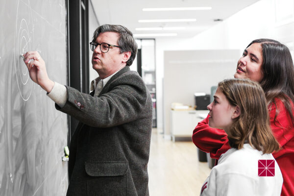 A professor writes on a blackboard while two students watch.