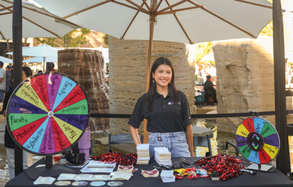 A smiling individual stands behind a table at an outdoor Chapman Fund booth, featuring colorful spinning wheels and promotional materials under a large red banner.
