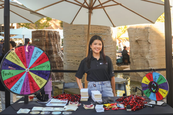 A smiling individual stands behind a table at an outdoor Chapman Fund booth, featuring colorful spinning wheels and promotional materials under a large red banner.