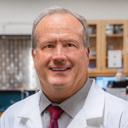 Headshot of Dean Rennolds Ostrom of CUSP, wearing a lab coat and smiling, set against a laboratory background filled with equipment.